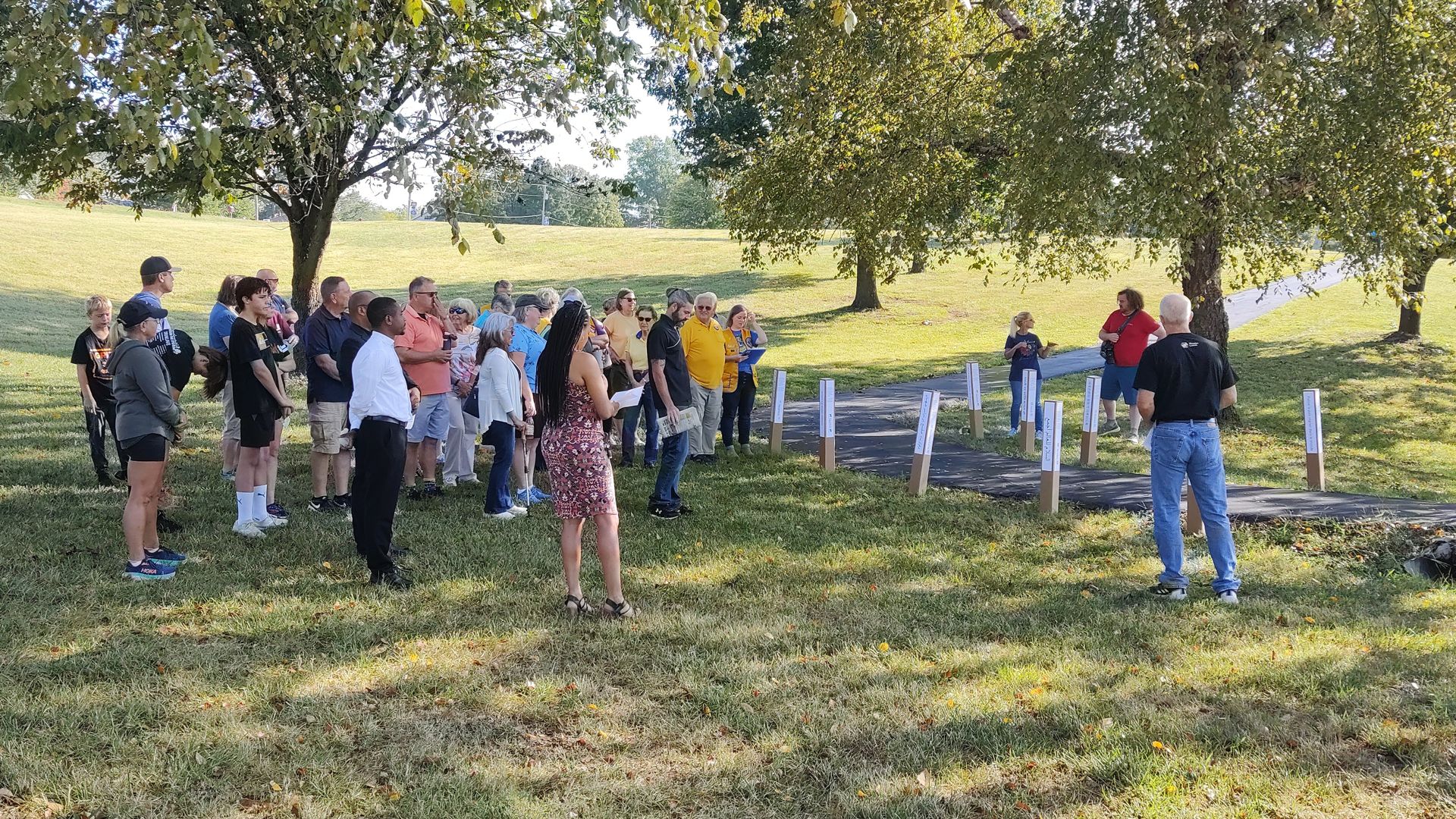 Approximately 35 people are gathered outside in Lakeview Park listening to speaker Paul Pecha from Waterloo Listens explain about Peace Poles.